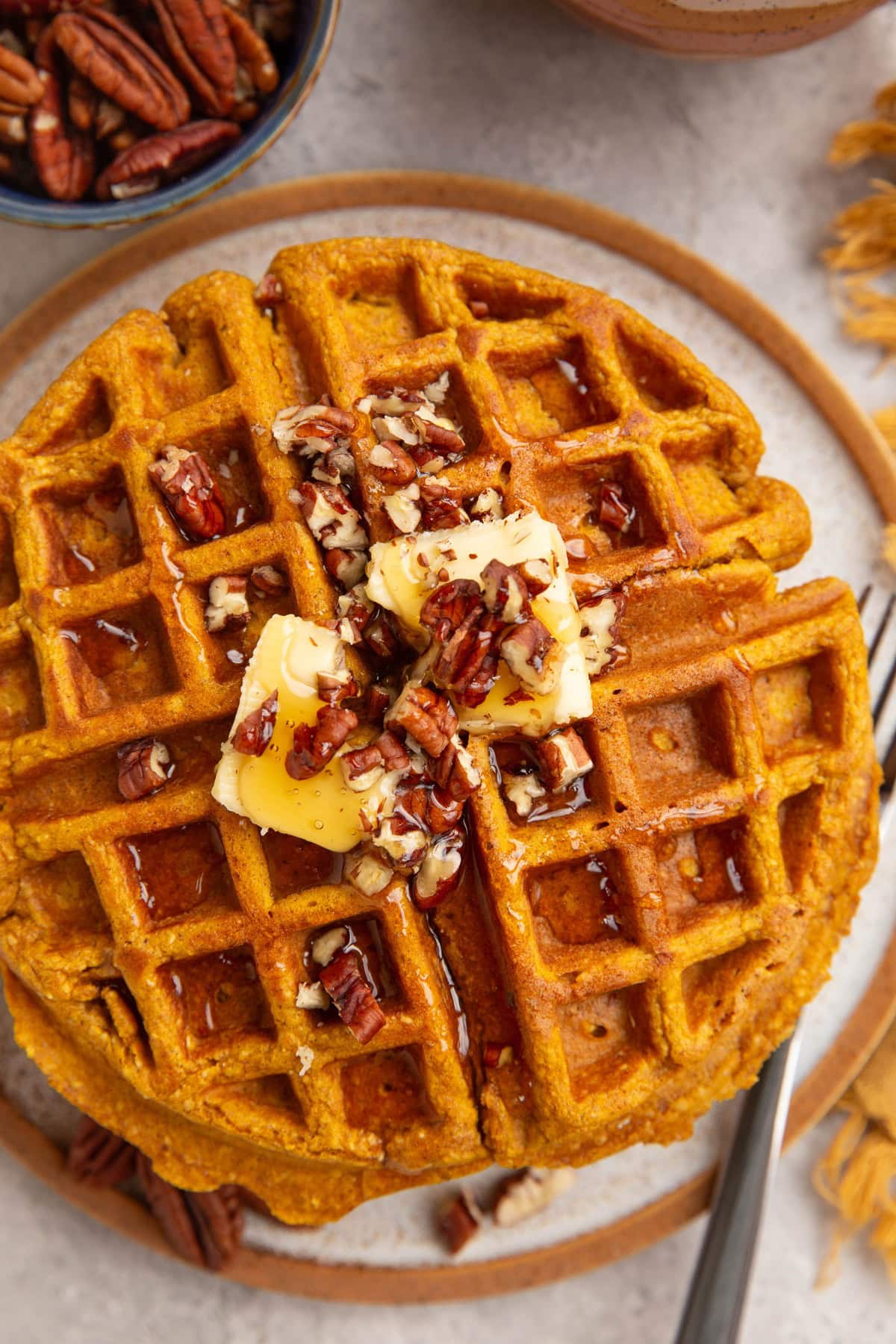 Pumpkin waffles on a plate, seen from the top down view with butter and chopped pecans on top and a fork to the side to serve.