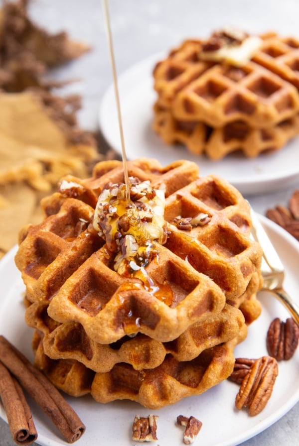 Drizzling honey over three sweet potato waffles with butter and chopped pecans on top. A plate of two waffles in the backfround.