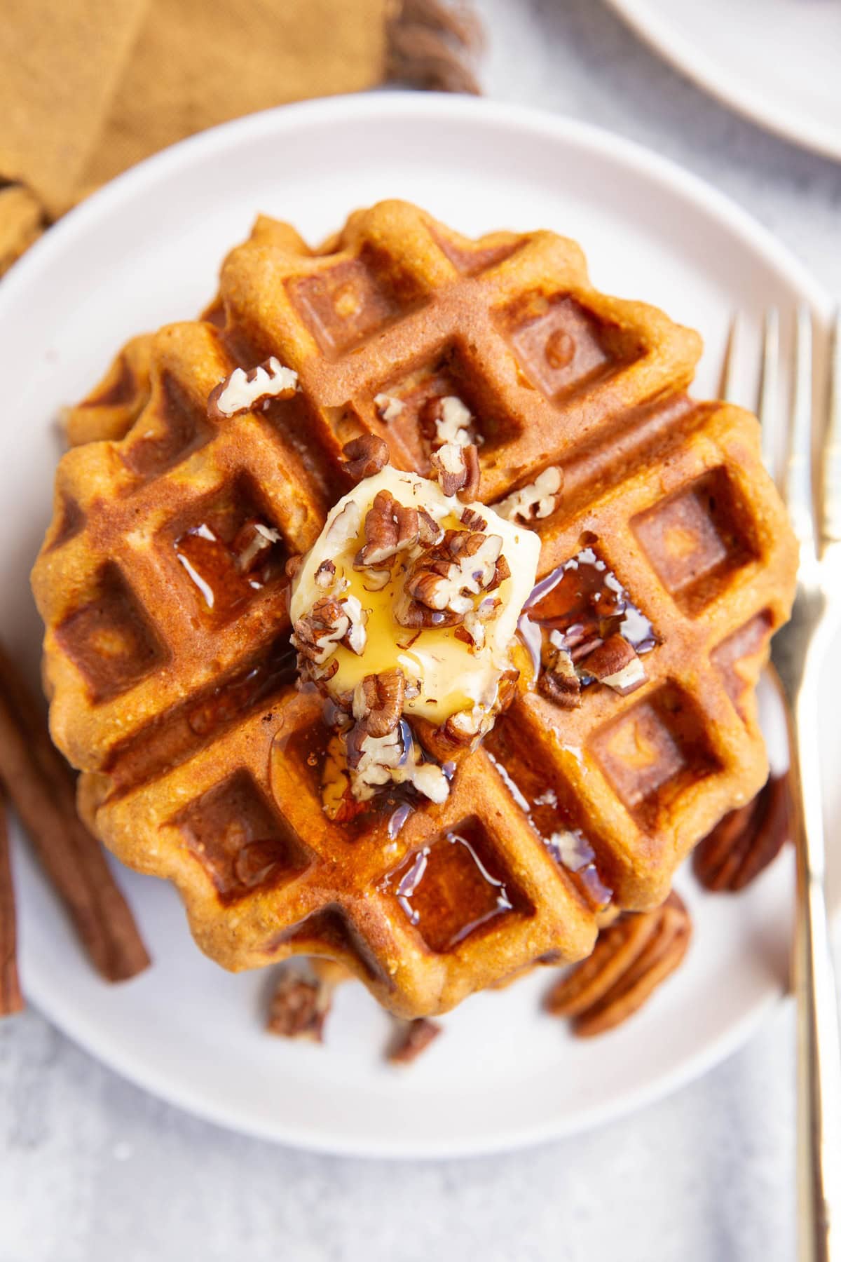 Sweet potato waffles on a white plate seen from the top view with butter, nuts and honey on top. A golden napkin and a golden fork to the side.