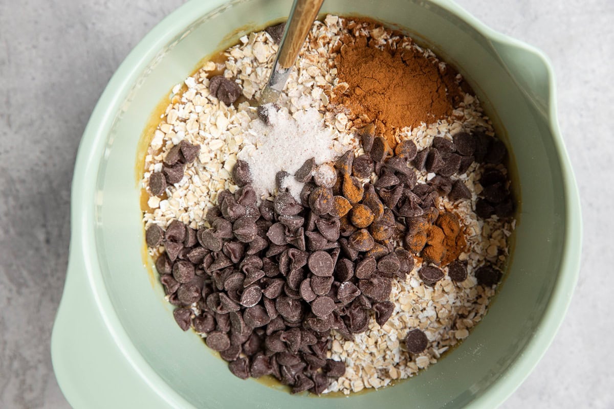 Mixing bowl with oats, chocolate chips, ground cinnamon, and salt on top of the wet ingredients, ready to be mixed in to make cookie dough.