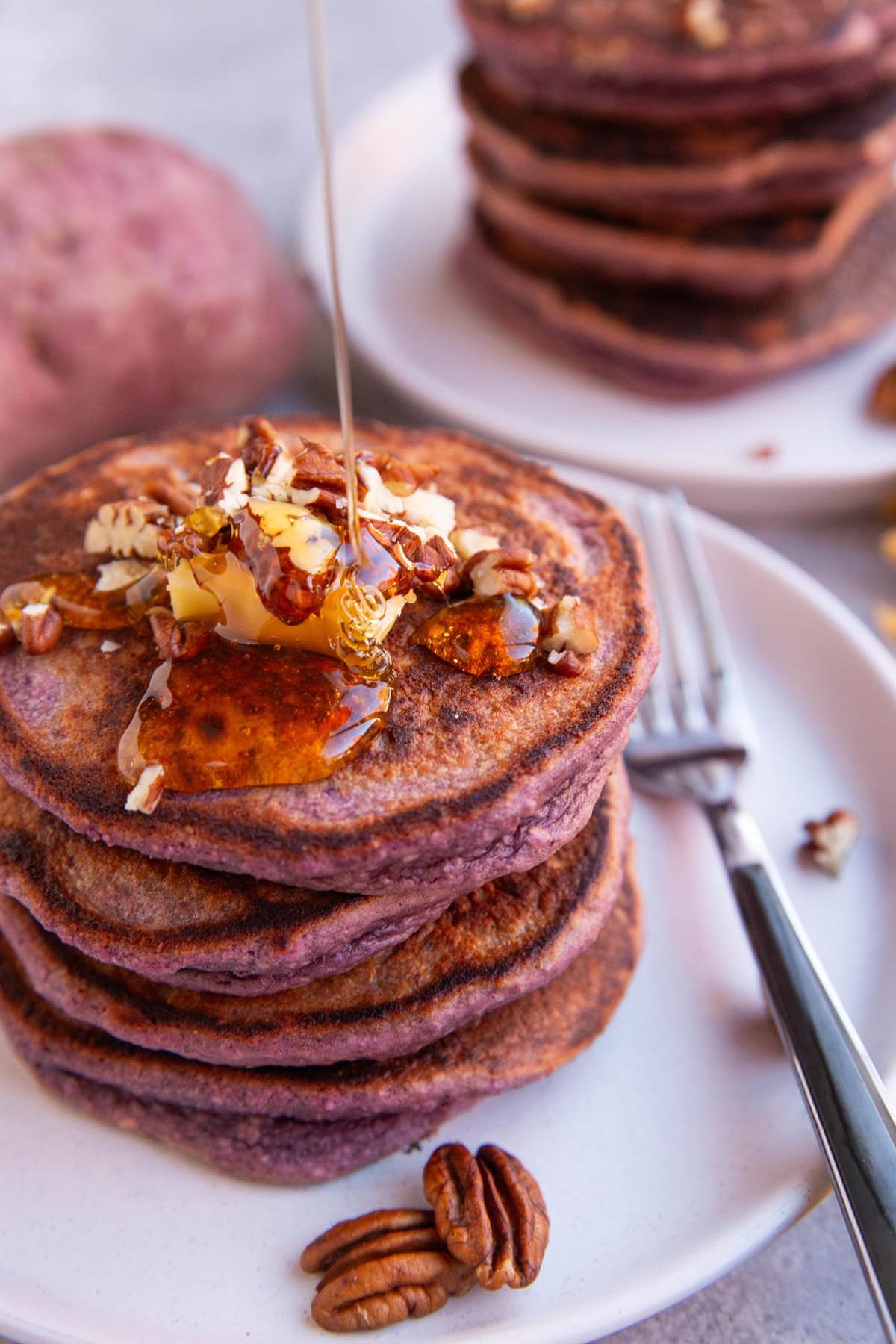 Drizzling honey over a stack of purple sweet potato pancakes with butter and chopped pecans.