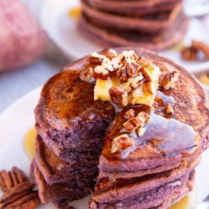 Stack of purple pancakes on a white plate with a bite taken out, topped with butter and chopped pecans.