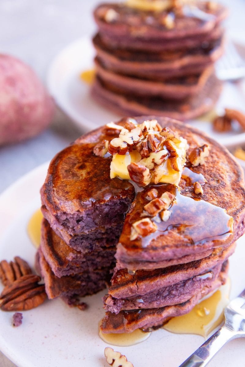 Stack of purple pancakes on a white plate with a bite taken out, topped with butter and chopped pecans.