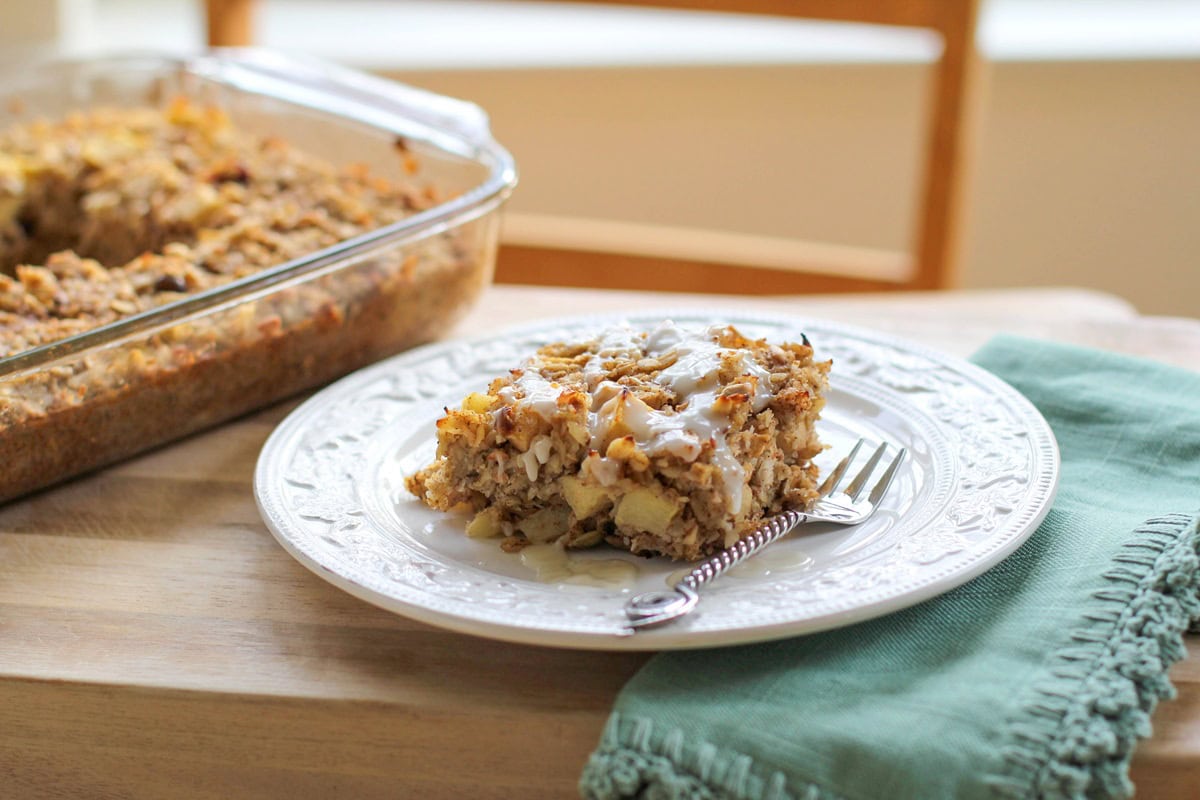 Slice of apple cinnamon baked oatmeal with a glaze drizzled on top, sitting on a white plate with the rest of the casserole dish full of oatmeal in the background.