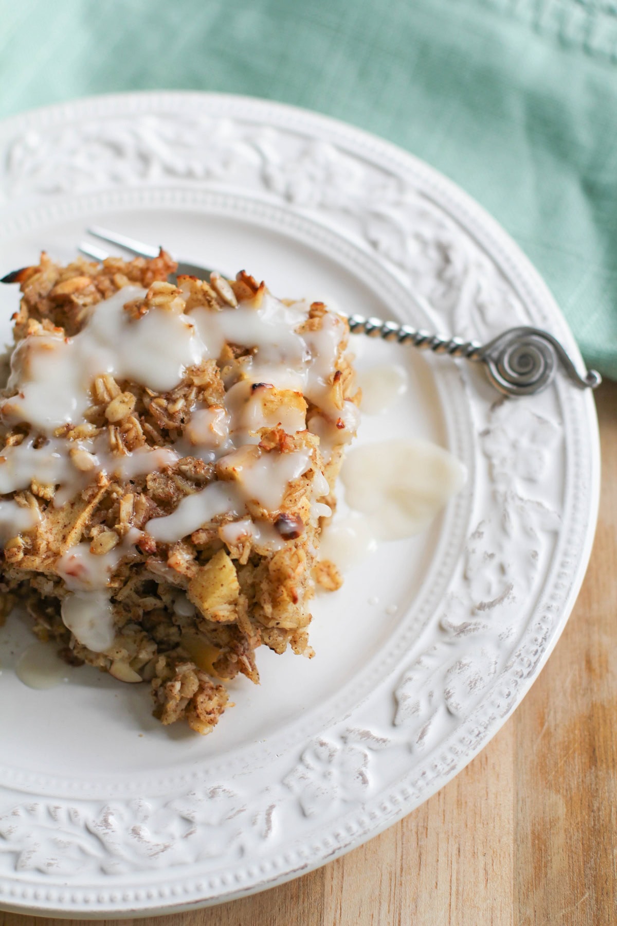 Apple baked oatmeal drizzled with glaze on a white plate with a small fork and a teal napkin to the side.