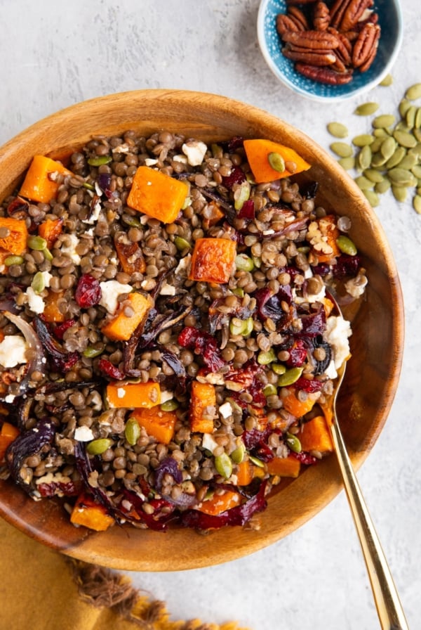 Wooden bowl full of butternut squash lentil salad with dried cranberries, pecans, pumpkin seeds, and maple balsamic vinaigrette. A golden napkin to the side, a small bowl full of pecans, and pumpkin seeds to the side. A golden spoon inside the bowl, ready to serve the salad