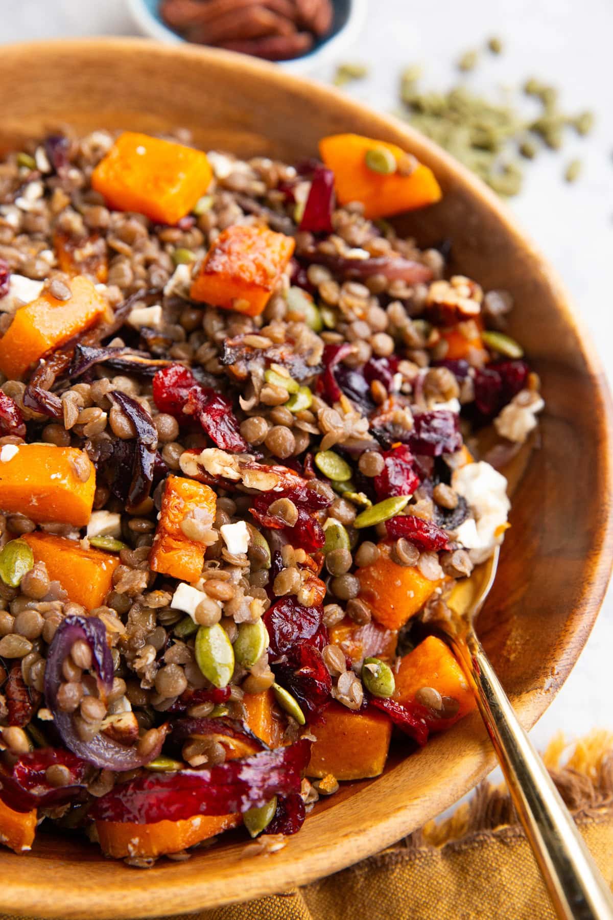 Butternut squash lentil salad in a large serving bowl with a golden spoon, ready to serve. Pecans and pumpkin seeds in the background.