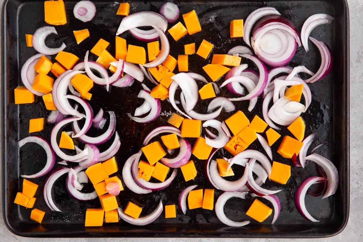 Baking sheet with chopped butternut squash cubes and sliced red onion tossed in oil and salt, ready to go into the oven.