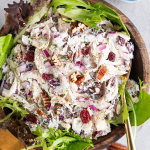 Turkey salad with fresh greens in a wooden bowl with a golden napkin and a small bowl of pecans to the side. A golden spoon is ready to serve the salad.