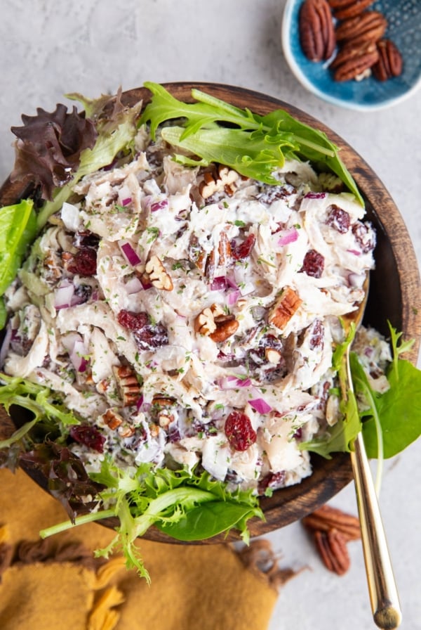 Turkey salad with fresh greens in a wooden bowl with a golden napkin and a small bowl of pecans to the side. A golden spoon is ready to serve the salad.