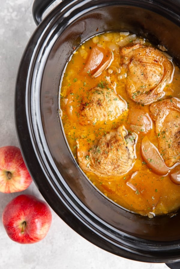 Slow cooker full of bone in chicken thighs, chopped apples, onion, and apple cider surrounding the cooked chicken.