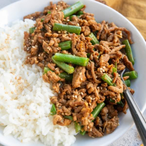 White bowl of white rice and garlic ginger ground turkey with green beans. a small blue bowl of sesame seeds in the background and some fresh cilantro in the foreground.