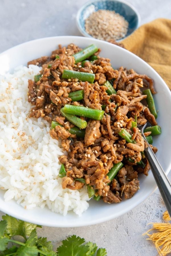 White bowl of white rice and garlic ginger ground turkey with green beans. a small blue bowl of sesame seeds in the background and some fresh cilantro in the foreground.