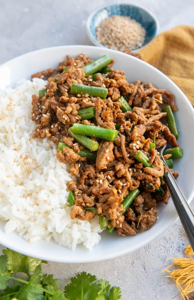 White bowl of white rice and garlic ginger ground turkey with green beans. a small blue bowl of sesame seeds in the background and some fresh cilantro in the foreground.