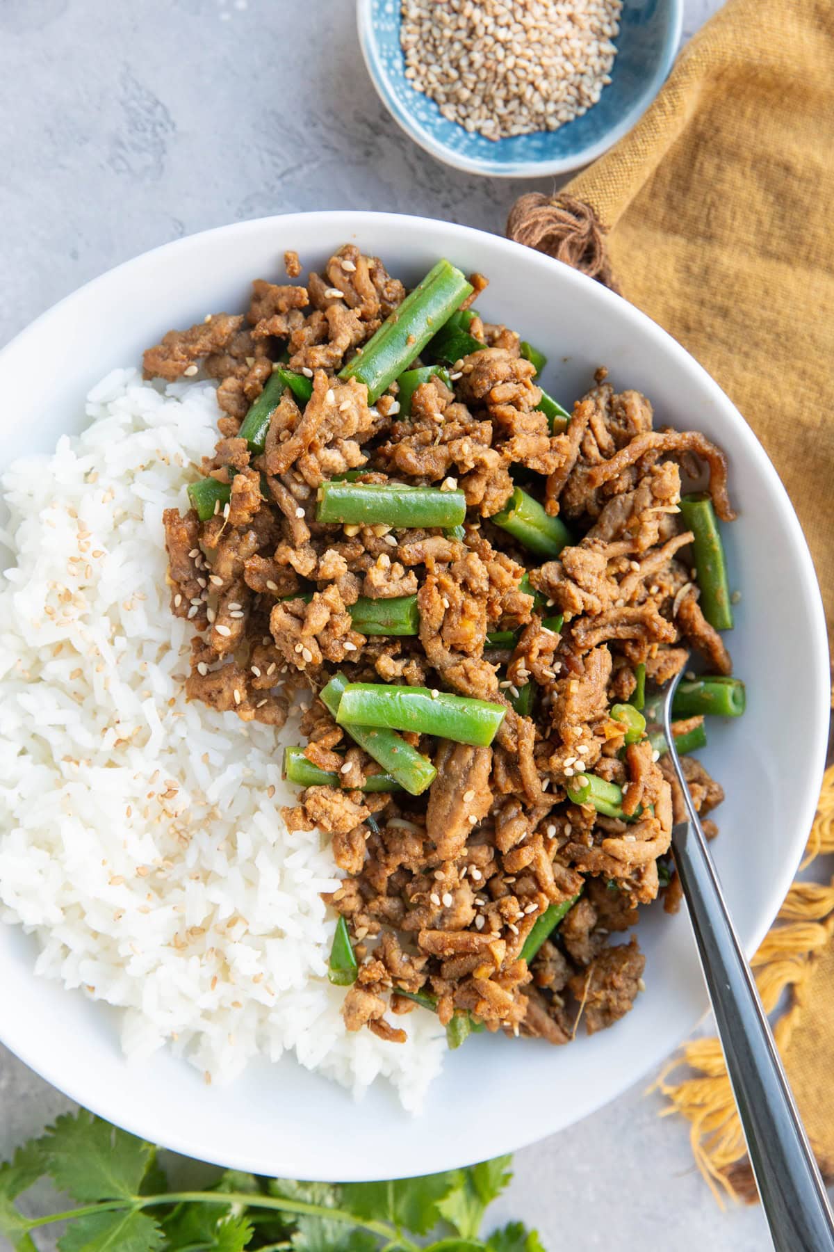 Garlic Ginger Ground Turkey recipe in a white bowl with white rice and a fork to the side, ready to eat. A small blue bowl holds sesame seeds, there's a golden napkin and fresh cilantro to the side.