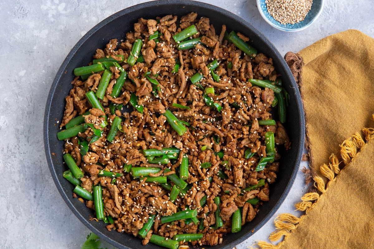 Skillet with garlic ginger ground turkey with green beans and a bowl of sesame seeds to the side with a golden napkin, ready to serve.
