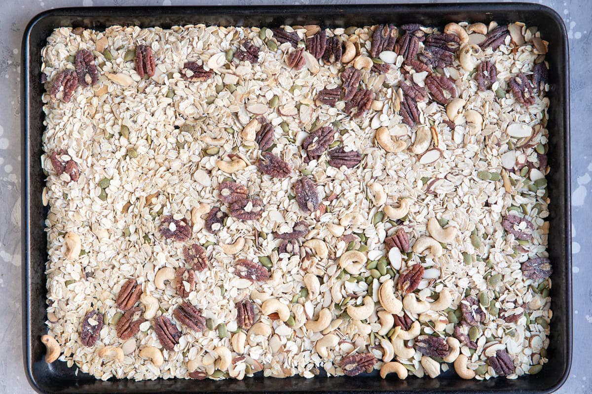 Large rimmed baking sheet with oats, cashews, sliced almonds, pecans, and pumpkin seeds spread over it.
