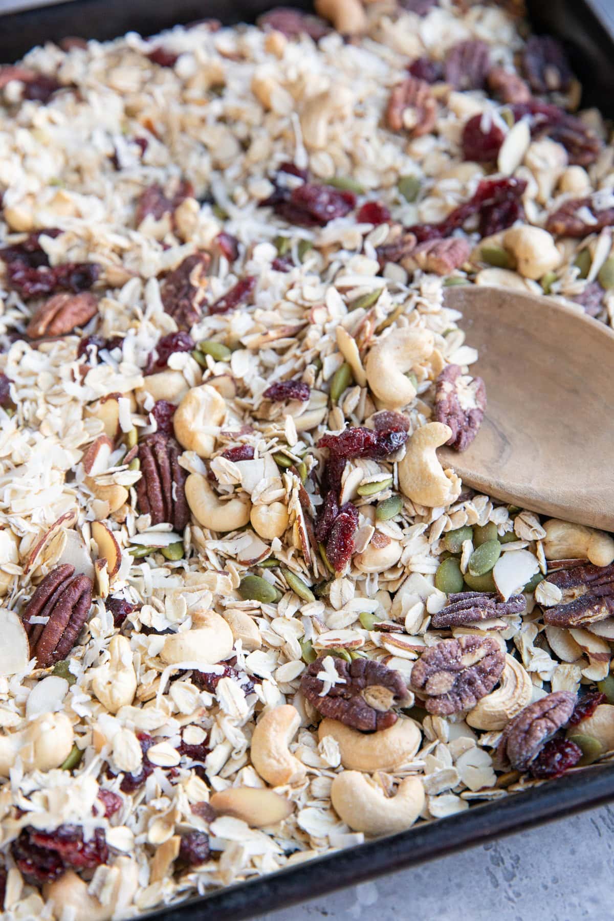 Homemade muesli on a large baking sheet with a wooden spoon scooping some of it.