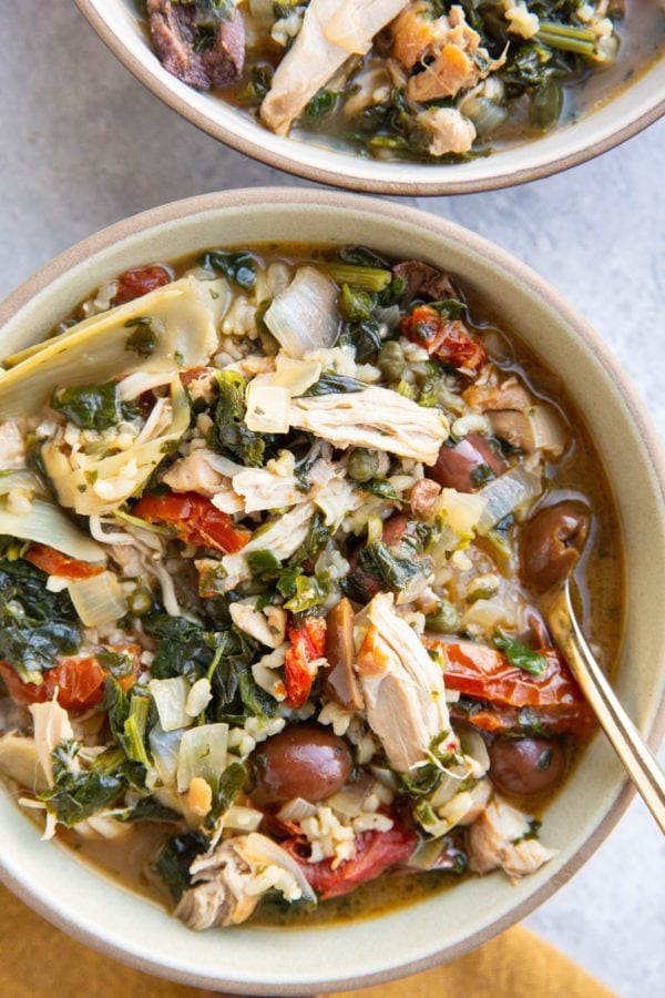 Mediterranean Chicken Soup with brown rice, Kalamata olives, sun-dried tomatoes and more in two bowls with a golden napkin.