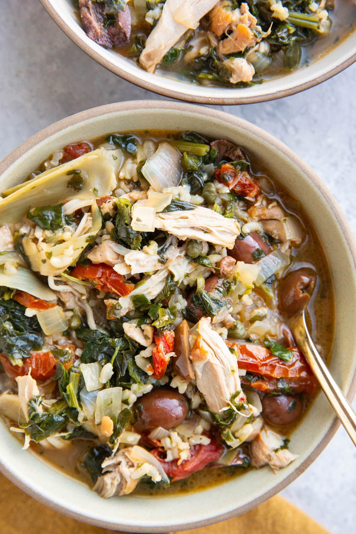 Mediterranean Chicken Soup with brown rice, Kalamata olives, sun-dried tomatoes and more in two bowls with a golden napkin.