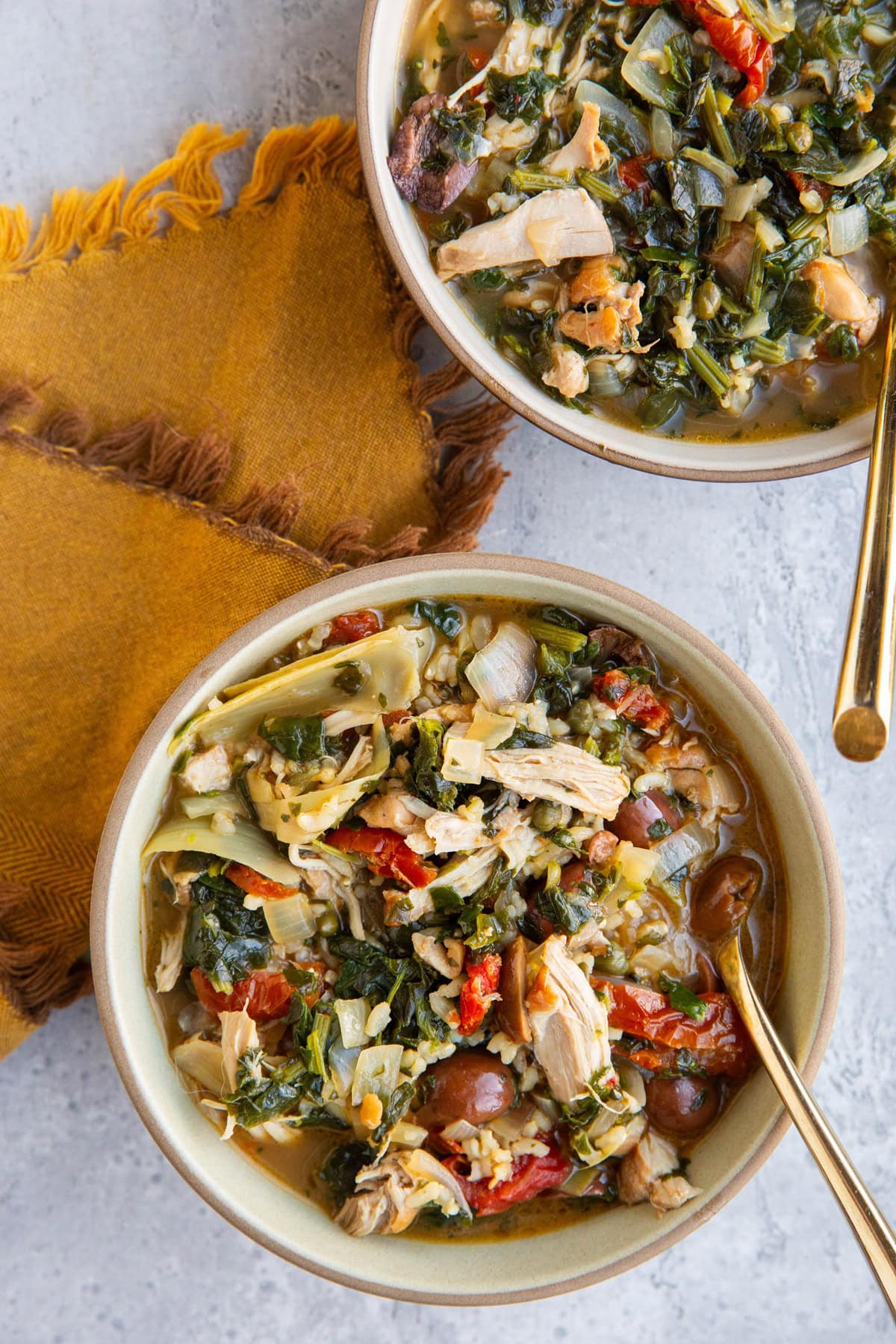 Two bowls full of Mediterranean Chicken Soup with a golden napkin and spoons, ready to serve.