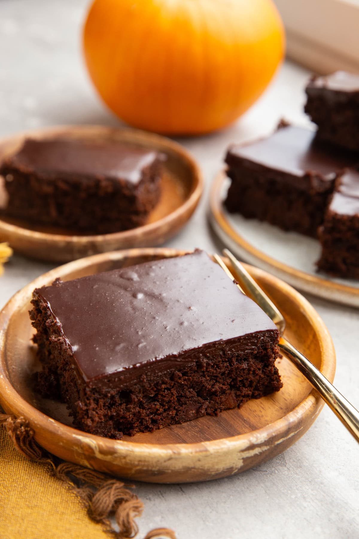Two slices of pumpkin chocolate cake on wooden plates with a plate with the rest of the cake in the background and a fresh pumpkin in the background.