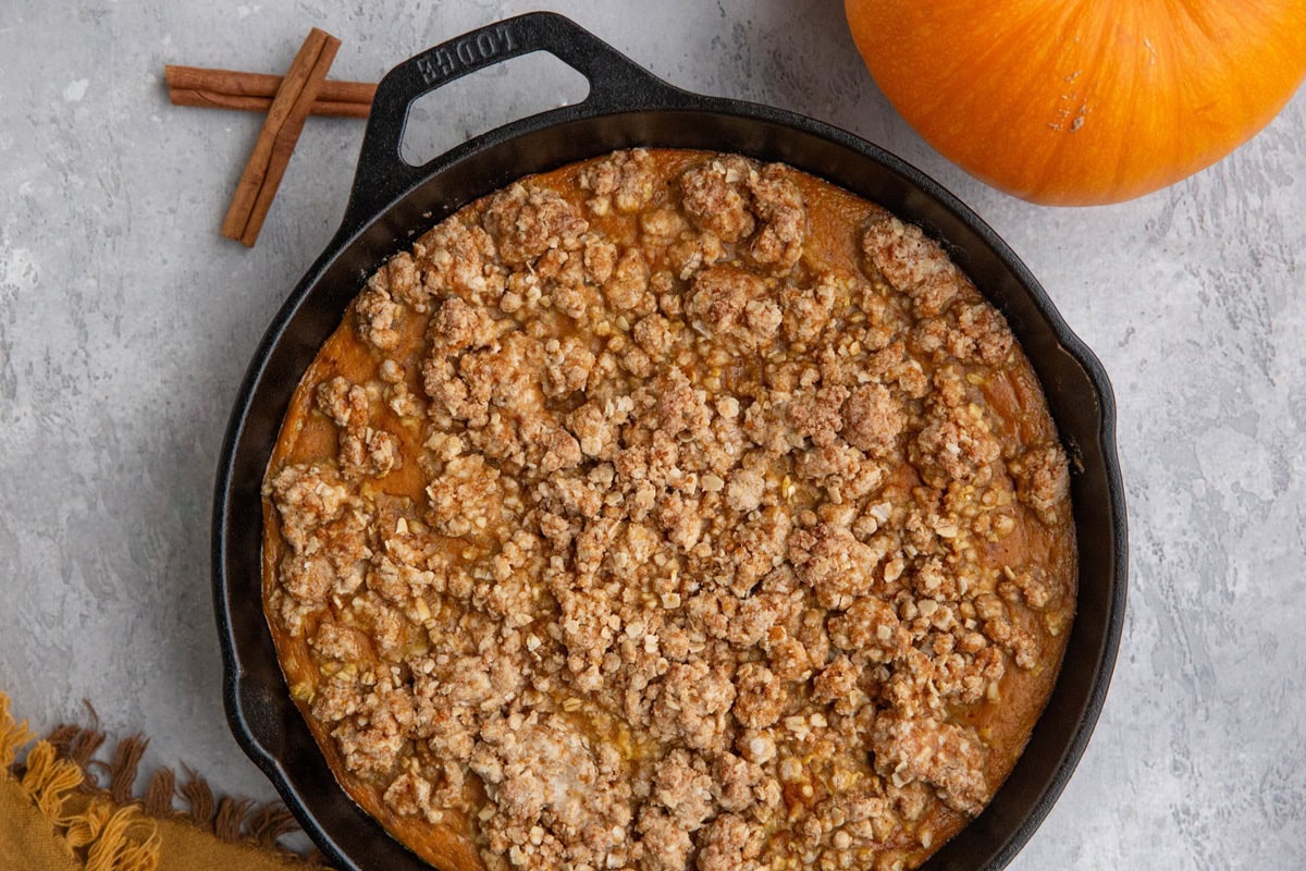 Cast iron skillet full of pumpkin crisp with an oatmeal streusel topping. A fresh pumpkin to the side and cinnamon sticks to the side.