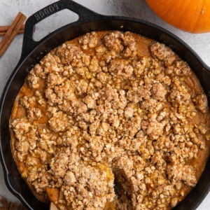 Cast iron skillet full of pumpkin crisp with a wooden spoon scooping out some of the crisp and a fresh pumpkin to the side.
