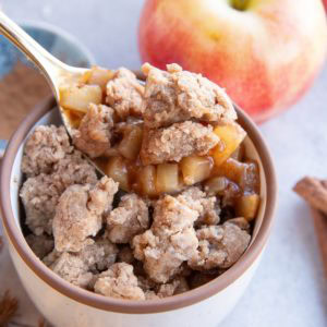 Ramekin with apple cobbler and fresh apples in the background, with a spoon scooping out some apple cobbler.