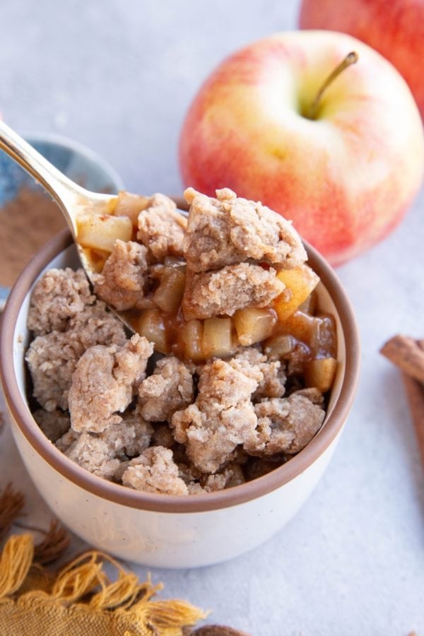 Ramekin with apple cobbler and fresh apples in the background, with a spoon scooping out some apple cobbler.