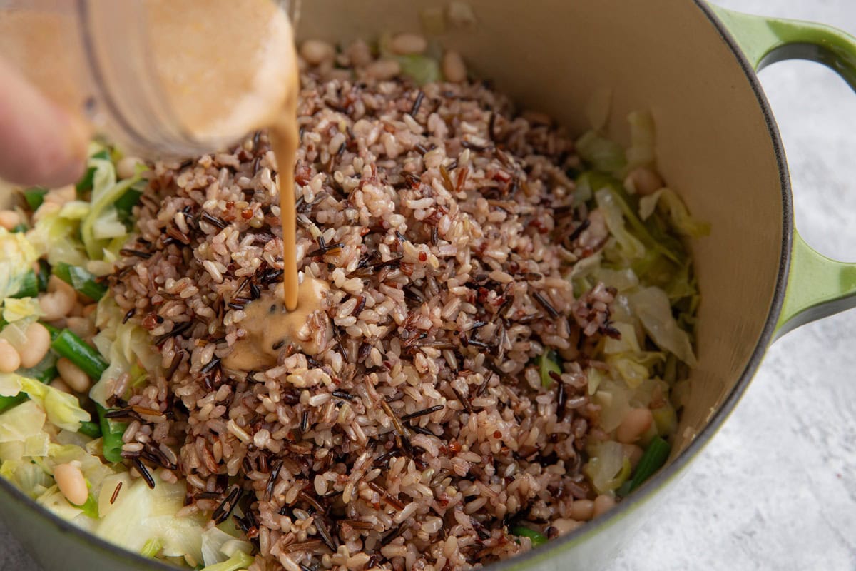 Large pot of vegetables with cooked wild rice on top with a hand pouring dressing in to make wild rice salad.