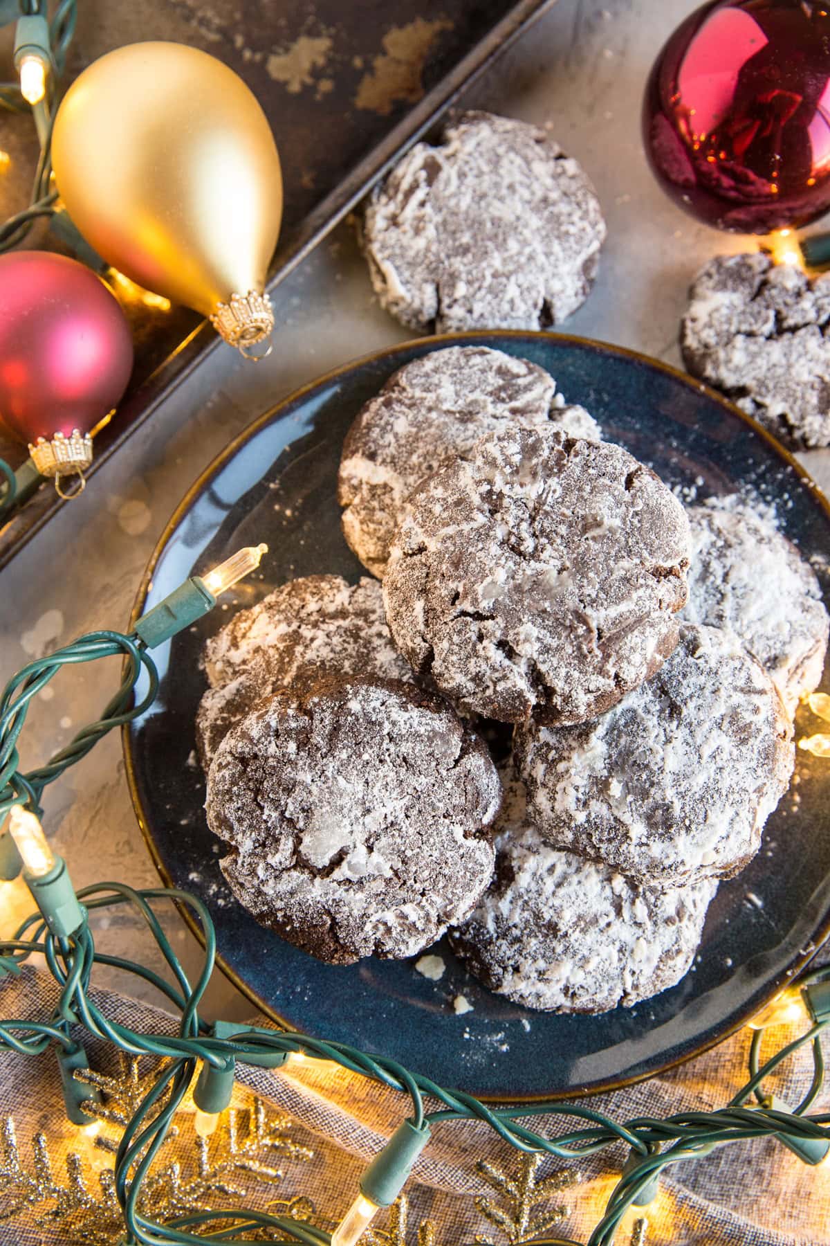 Blue plate filled with chocolate crinkle cookies covered with powdered sugar with Christmas likes and ornaments around the cookies.