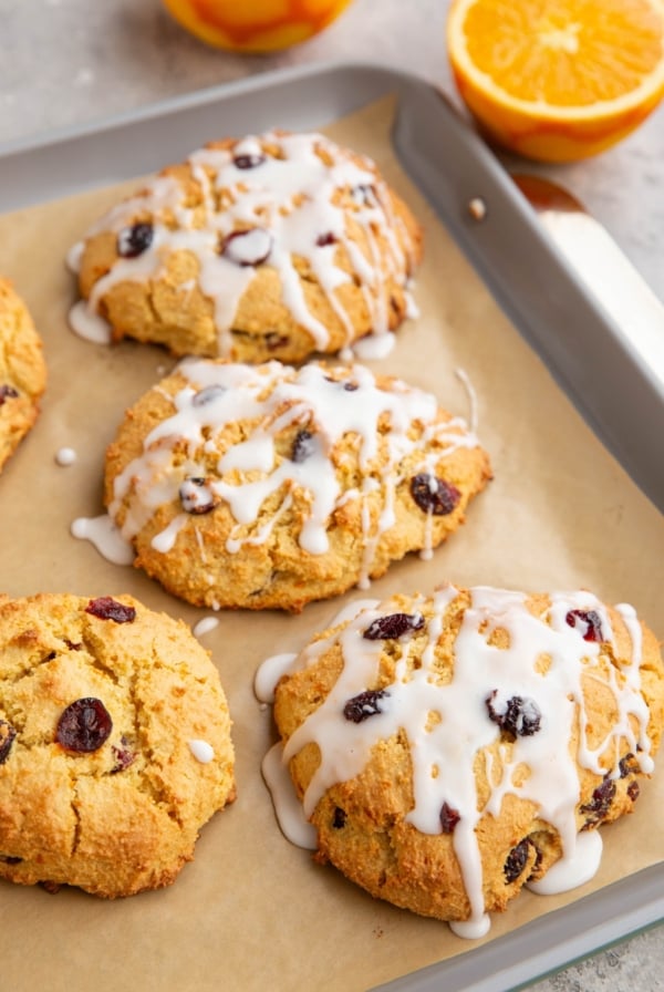 Baking sheet of cranberry orange scones drizzled with a glaze and a fresh orange in the background sliced in half.