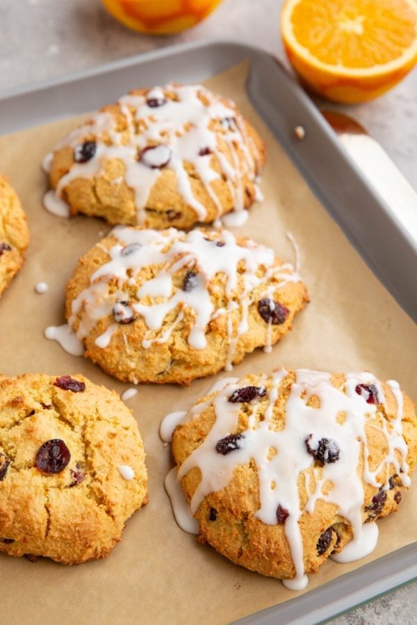 Baking sheet of cranberry orange scones drizzled with a glaze and a fresh orange in the background sliced in half.