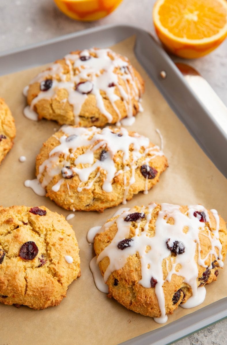 Baking sheet of cranberry orange scones drizzled with a glaze and a fresh orange in the background sliced in half.
