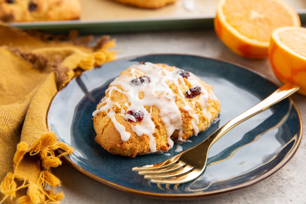 Cranberry orange scone on a blue plate with a gold fork and the rest of the scones and a fresh orange in the background.