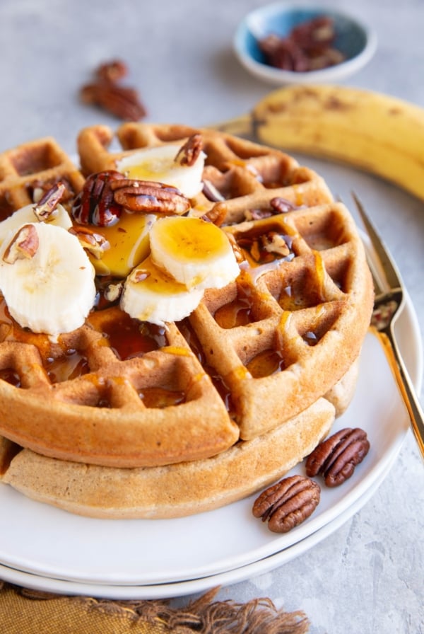 Two large Banana Oat Waffles on a white plate with sliced bananas, pecans and honey on top. A ripe banana and pecans in the background.