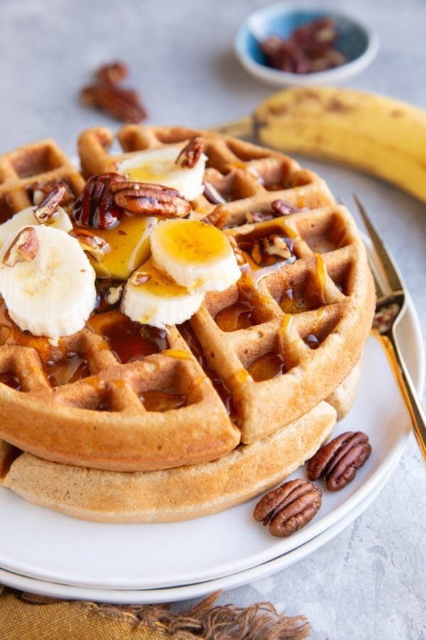 Two large Banana Oat Waffles on a white plate with sliced bananas, pecans and honey on top. A ripe banana and pecans in the background.