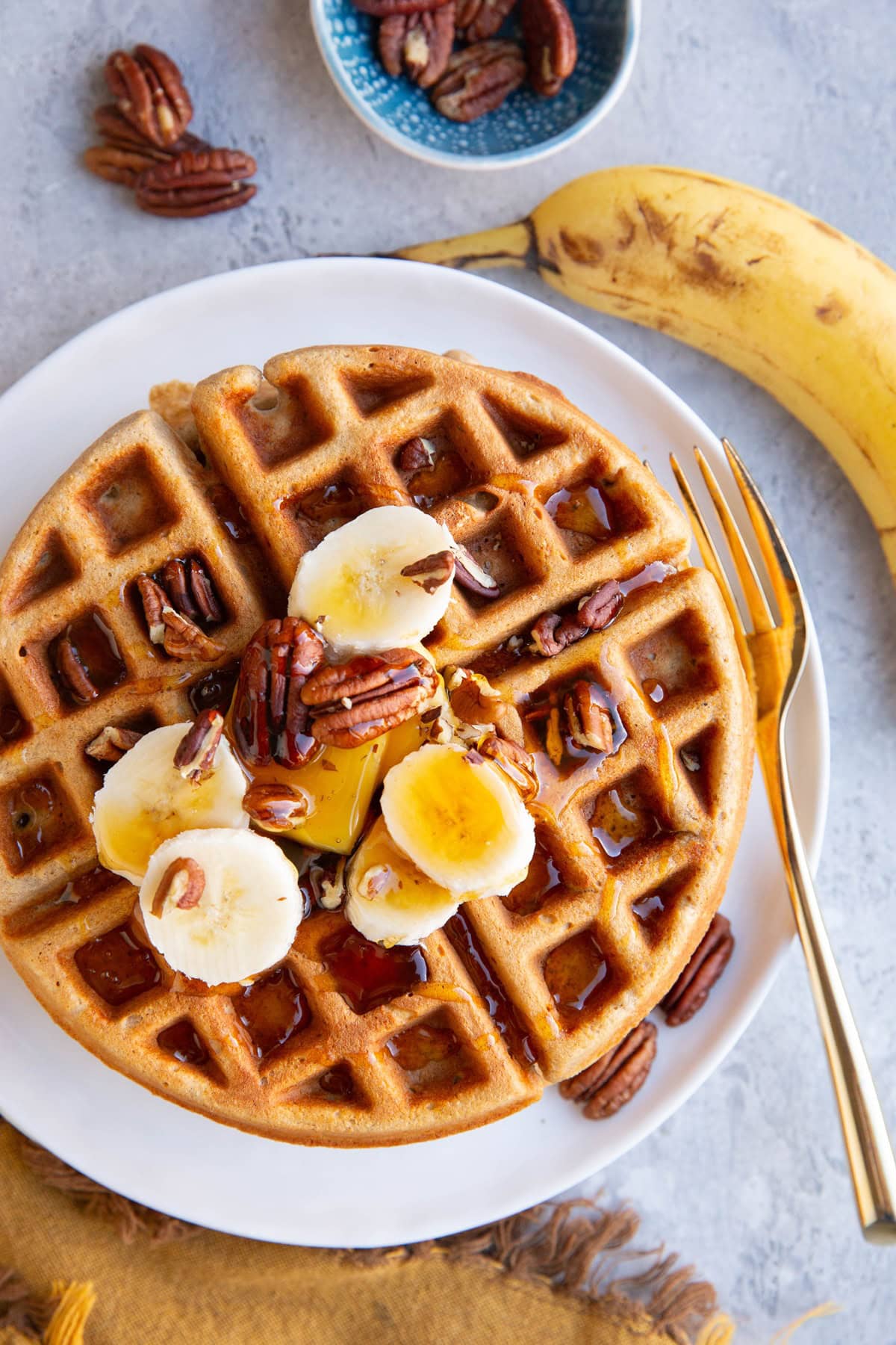 Banana oatmeal waffles on a white plate as seen from the top down with a gold fork to the side, ready to serve. A ripe banana and pecans to the side.