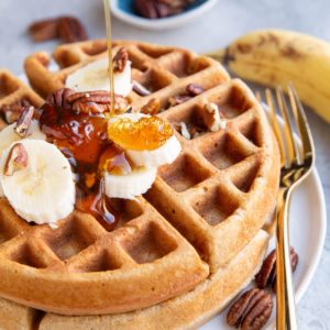 Honey being poured on top of banana oat waffles with sliced bananas and pecans on top. A golden fork to the side and a ripe banana and pecans in the background.