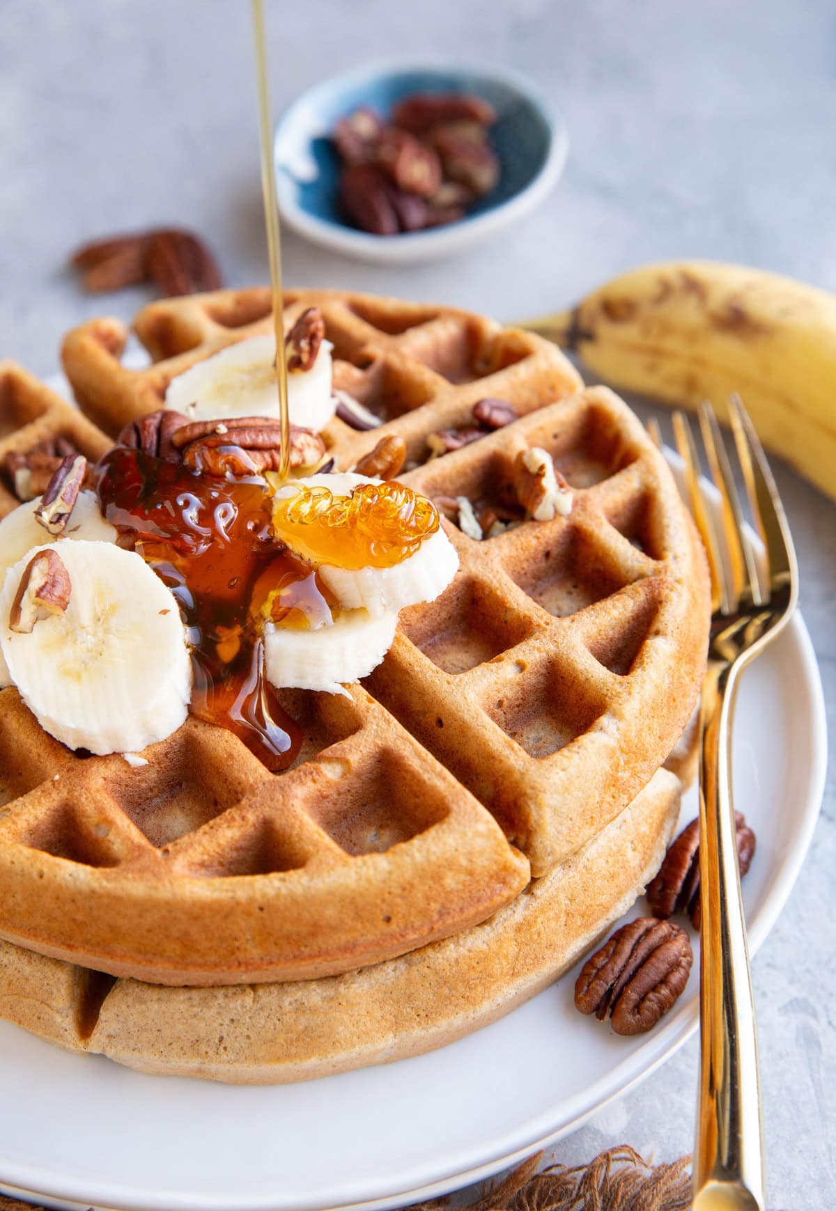 Honey being poured on top of banana oat waffles with sliced bananas and pecans on top. A golden fork to the side and a ripe banana and pecans in the background.