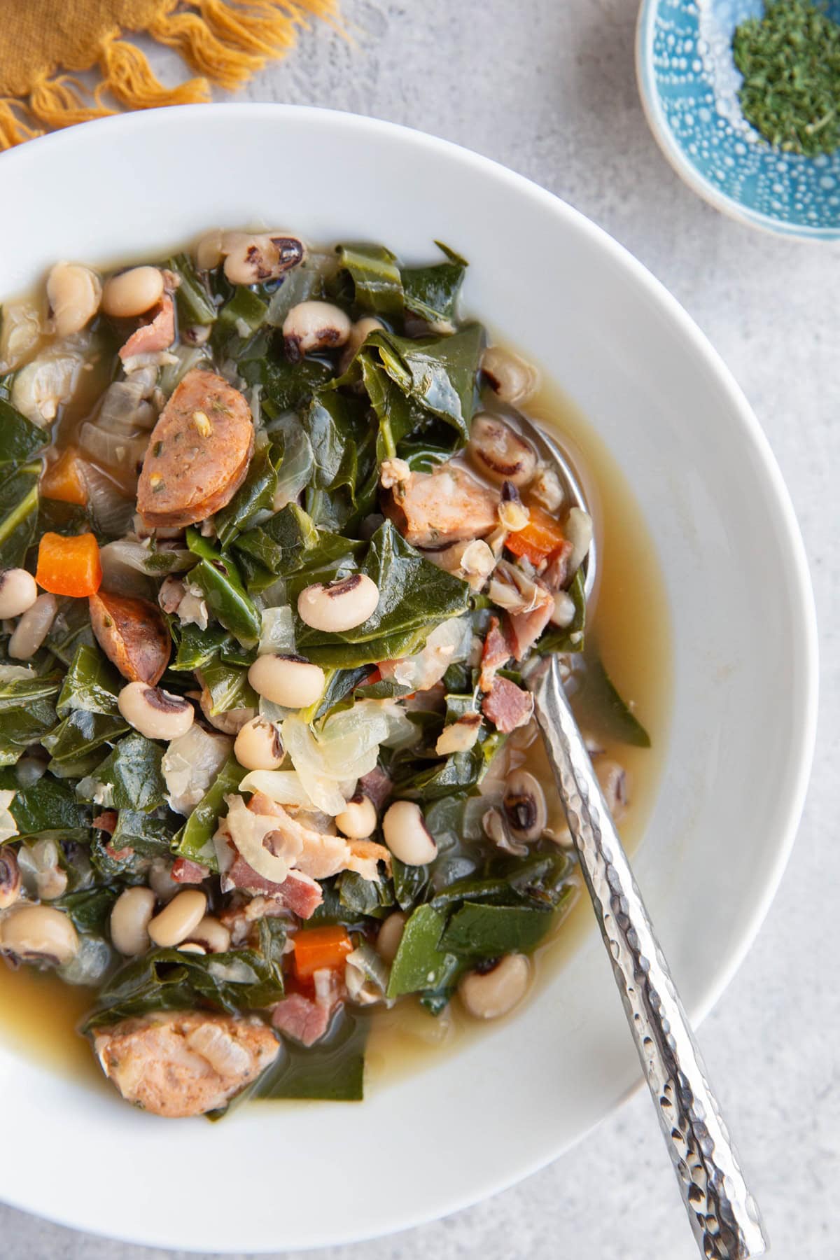 A big bowl of black eyed pea soup with collard greens, sausage, and a spoon on the side, ready to eat.