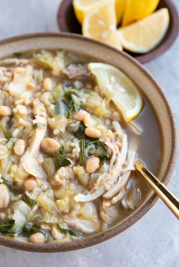 Large bowl of chicken, cabbage and white bean soup with spinach and lemon. A gold spoon inside and a bowl of lemon wedges.