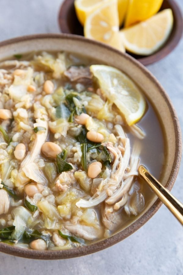 Large bowl of chicken, cabbage and white bean soup with spinach and lemon. A gold spoon inside and a bowl of lemon wedges.
