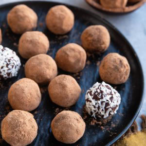 Black plate full of chocolate truffles that are coated with cocoa powder and two are covered with shredded coconut. Ready to serve.
