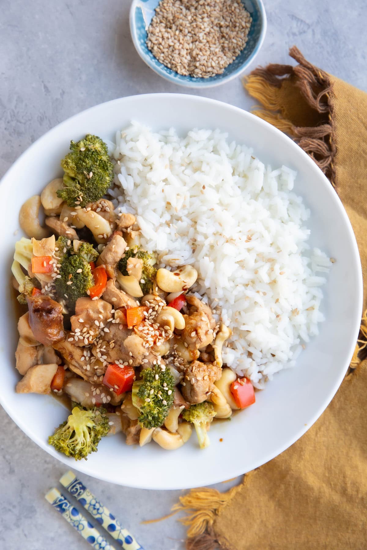 White bowl full of white rice and cashew chicken. A golden napkin, chopsticks, and a bowl of sesame seeds to the side.