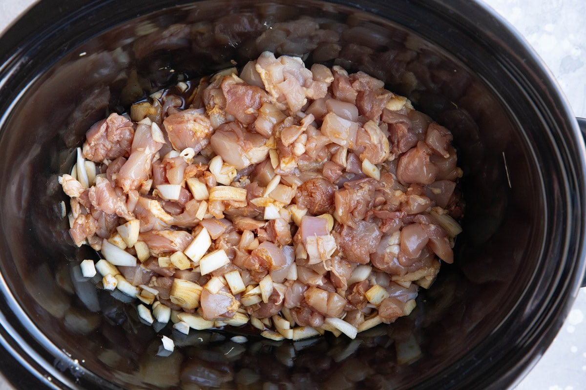 Slow cooker full of chopped chicken thighs, onions, sauce, and garlic.
