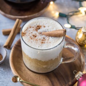 Eggnog latte in a clear coffee mug with a Chemex full of coffee in the background and Christmas lights and ornaments around.