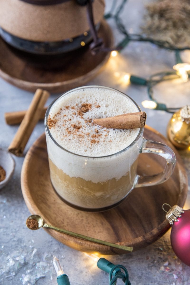 Eggnog latte in a clear coffee mug with a Chemex full of coffee in the background and Christmas lights and ornaments around.