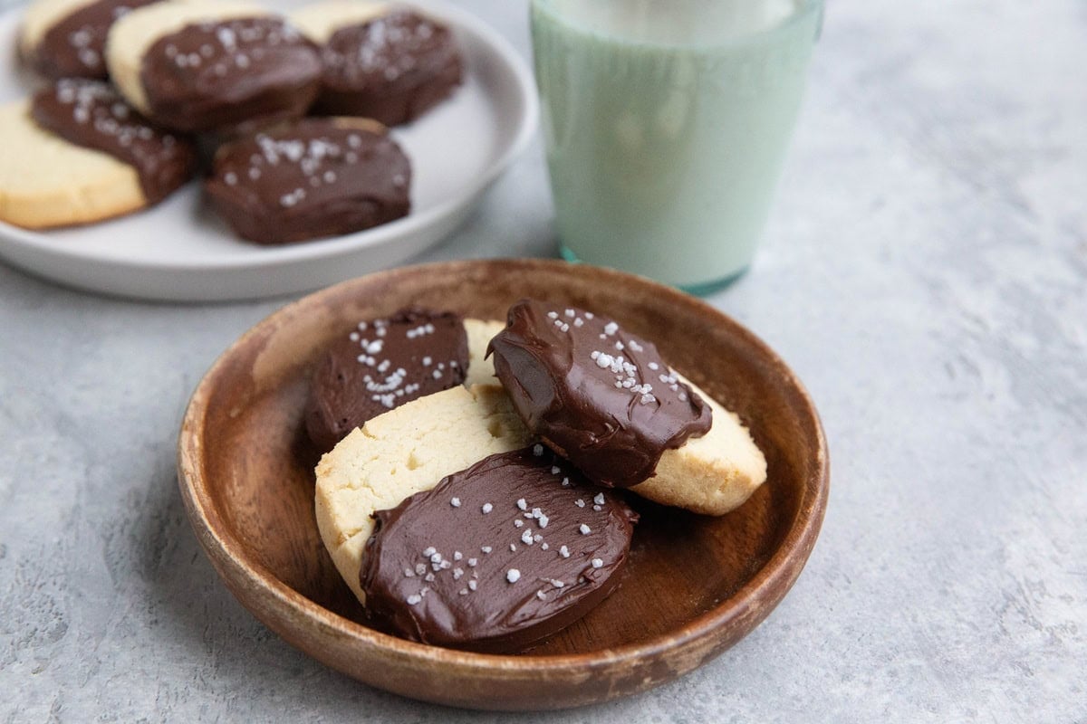 Wooden plate of chocolate dipped shortbread cookies sprinkled with flaky sea salt. A white plate of more cookies and a glass of milk in the background.